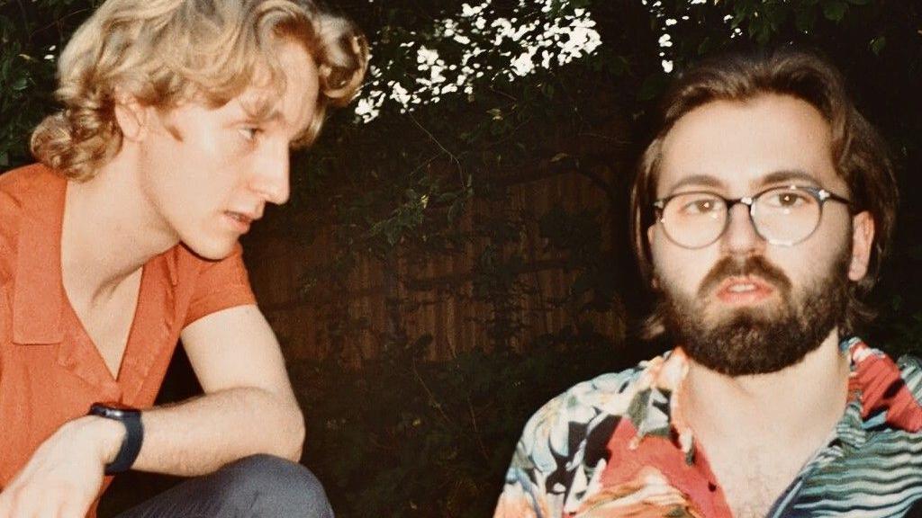 Two men, one is blond with an orange polo on, while the other has brown hair with a beard and glasses.
