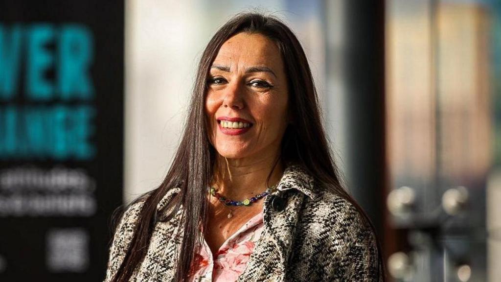 A photo of Eva Komuves smiling at the camera. She has long brown hair and is wearing a tweed black, white and yellow patterned jacket with a floral pink top underneath. She is also wearing a multi-coloured necklace. She is standing by a blurred out sign.