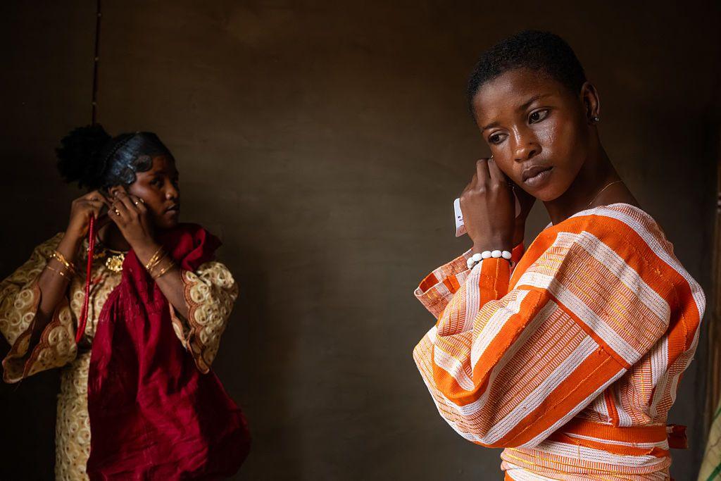 Two women put on earrings as they get ready. One is in an orange and white stripped outfit the other is in a cream and maroon dress - Friday 31 October 2025.