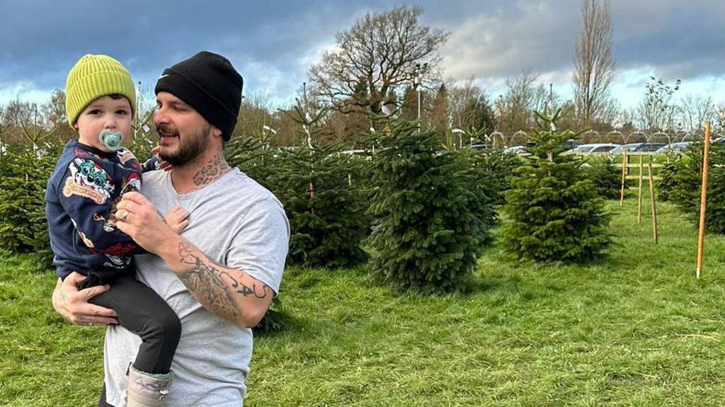 A man wearing a black beanie and a light grey t-shirt is holding his young son, who is wearing a blue sweater with a pattern on it, a green beanie and has a blue dummy in his mouth. The man has tattoos on his neck and arms. There are rows of Christmas trees behind them