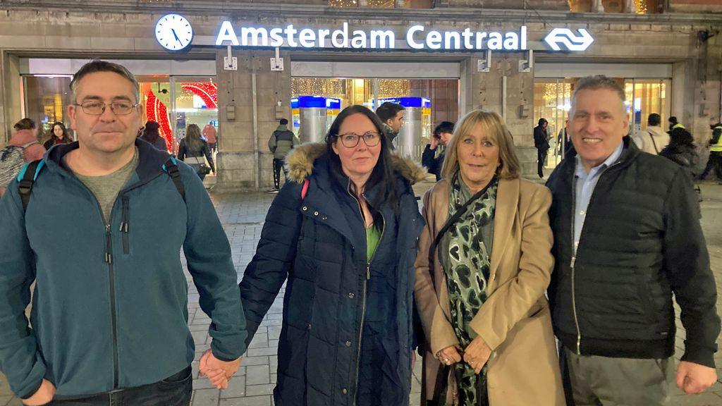 Four adults standing outside Amsterdam Central station. The man on the left, who is wearing a fleece, a rucksack and glasses, holds hands with his wife. She has long dark hair, glasses and a navy padded coat with a fur-lined hood. Next to her stands a woman with blonde, shoulder-length hair, wearing a trench coat and a patterned scarf. Next to her is a man with a shirt and chinos, a black jacket and short grey hair. The station's sign can be seen above the entrance behind them.