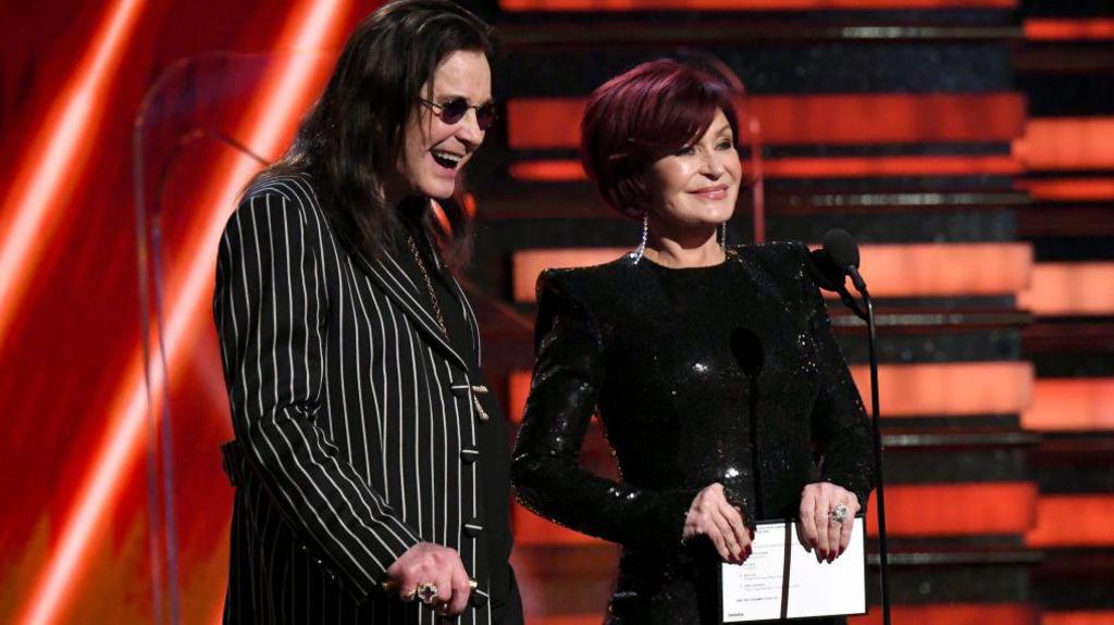 Ozzy Osbourne and Sharon Osbourne on stage at the Grammy awards in 2020. He is wearing a striped jacket and she has got an elegant black dress on. 