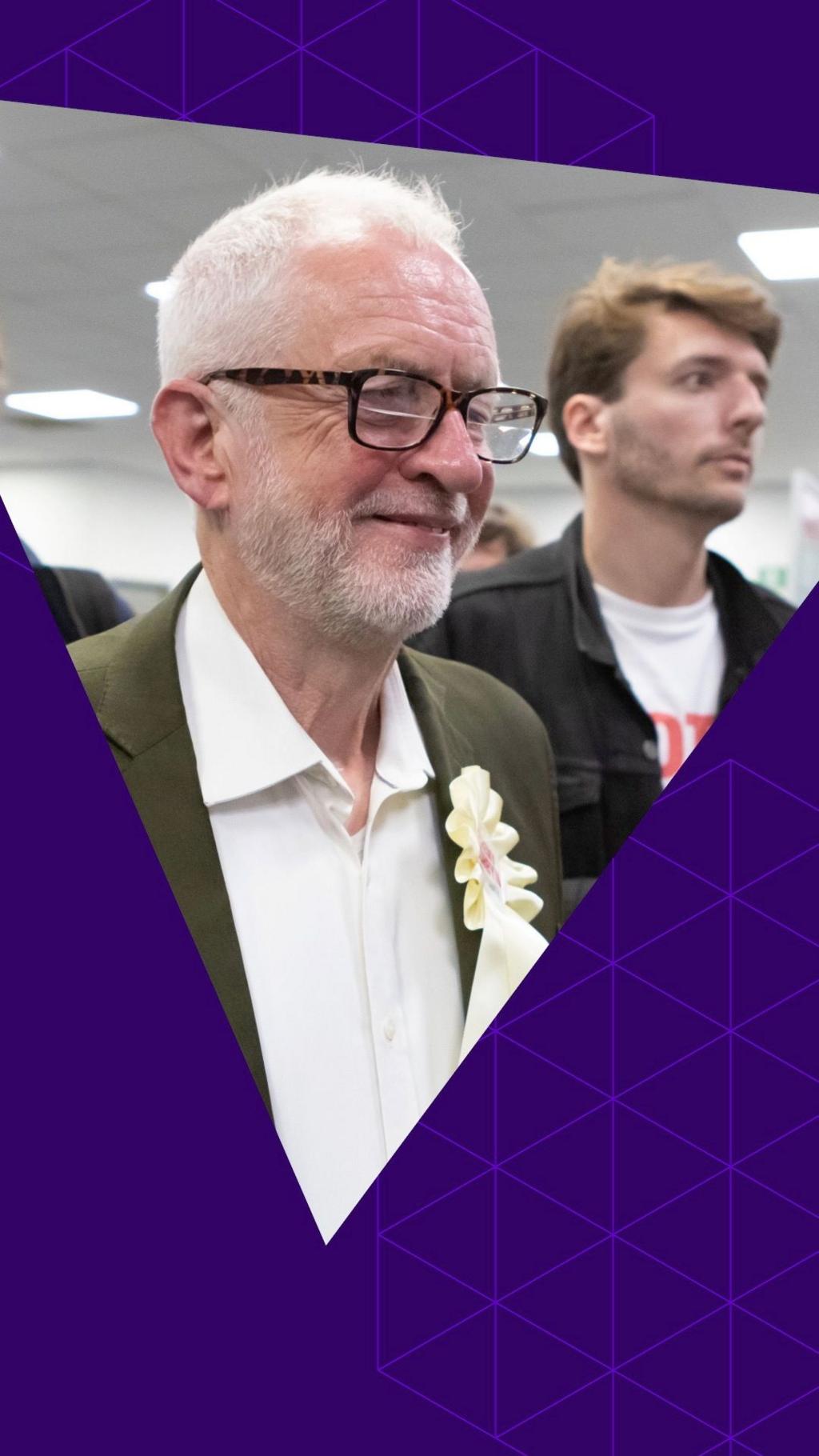 Jeremy Corbyn smiles. He is wearing dark rimmed glasses and a green suit jacket with a white shirt 