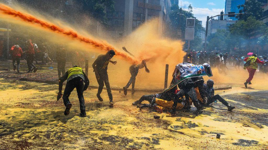 A streak of yellow-dyed water aimed at protesters comes from the left of the picture and hits the back of one man. Others try to avoid the spray and appear piled on the road surface on the right, ducking for cover.