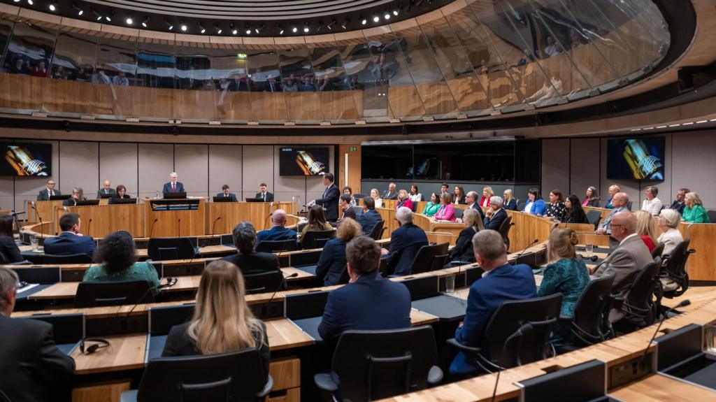 People sit in sits in the Senedd chamber, a hemicircle of brown desks with glass panelling surrounding them in a full circle from above.