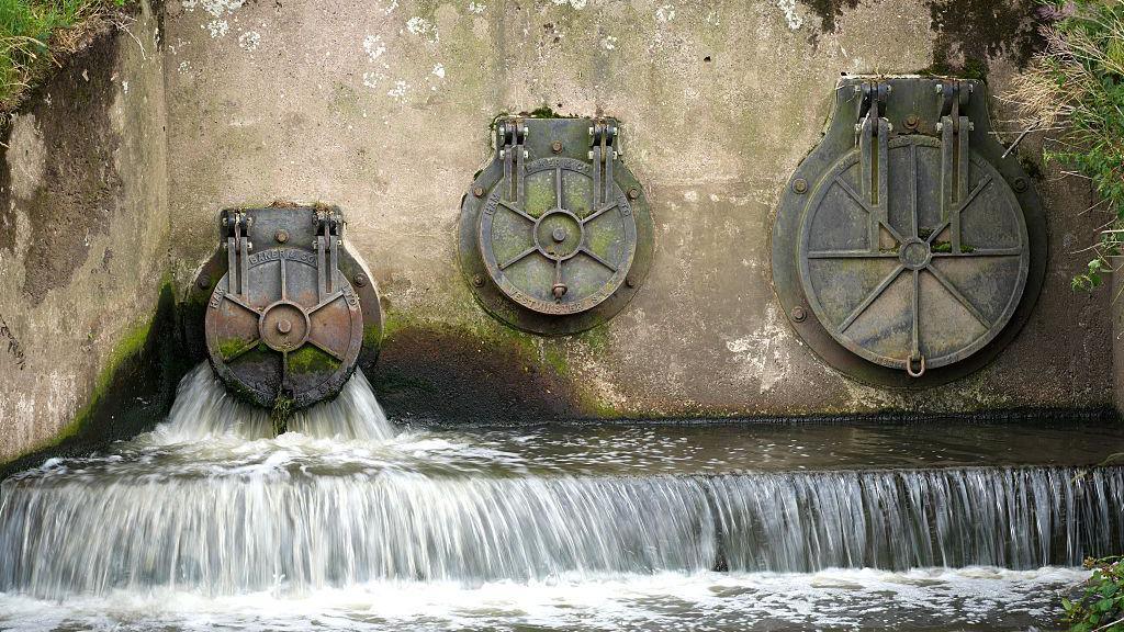 Water discharges from an outlet pipe. There are three pipes shown on a concrete wall with some moss visible.