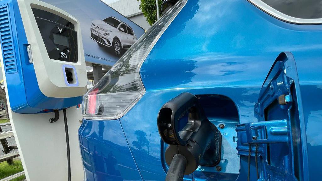 A bright blue VinFast electric car plugged in at an outdoor charging station. Only the rear of the vehicle can be seen, with the charging hatch open. A picture of a grey version of the vehicle can be seen next to the car.