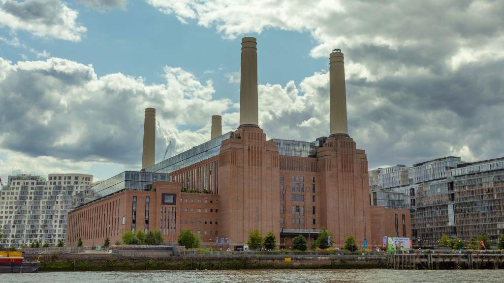 Battersea Power Station, a large red brick building on the edge of the Thames. It has a high chimney in each corner and is surrounded by low-level greenery. Office blocks can be seen around it.