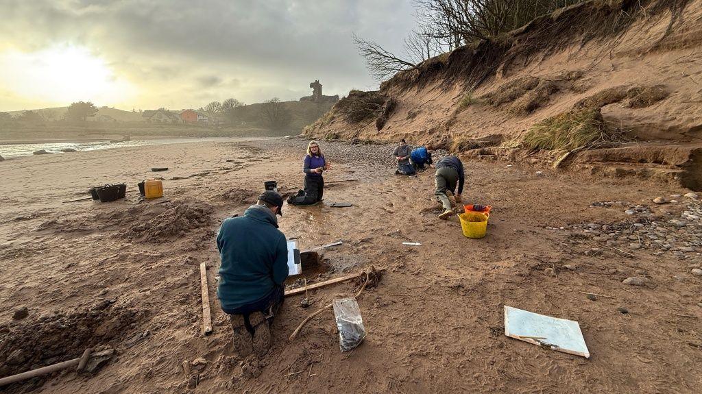 Five archaeologists kneel on the beach as they study the footprints. They're wearing wellies and are surrounded by tools and buckets. The sky is cloudy.