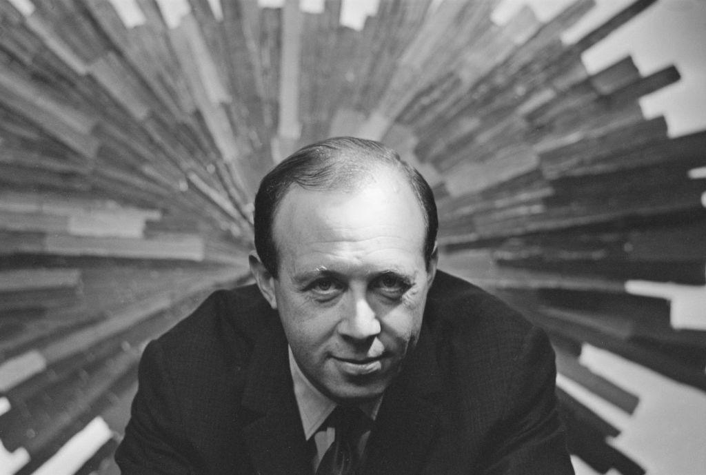 A black and white image of the late Bernat Klein - a balding man in a suit and tie with a large design in the background