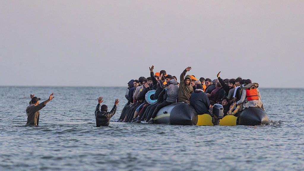 Two men standing in waist high water wave to an inflatable boat full of migrants attempting to cross the English Channel.
