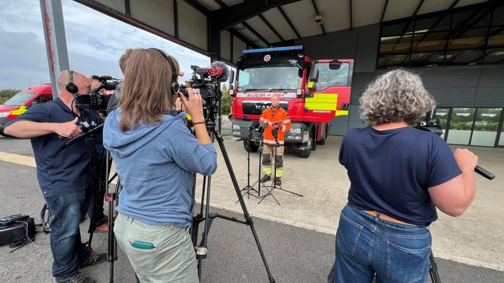 A man wearing an orange hi-vis jacket addresses the media