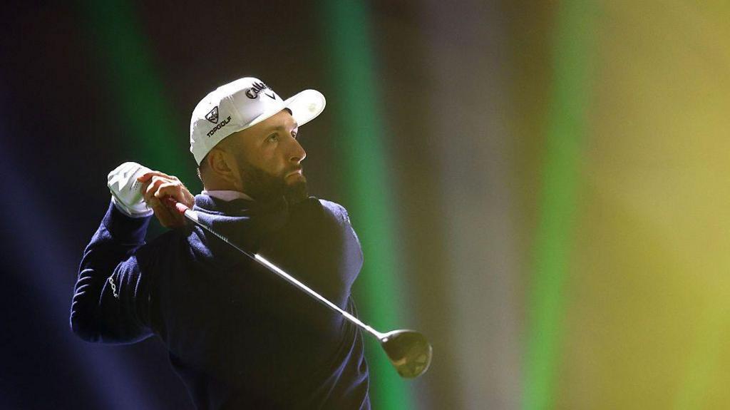 Jon Rahm tees off on the 16th hole on day one of LIV Golf Riyadh. He has a black beard and is wearing a white cap, a white glove on his left hand and a navy sweater.
