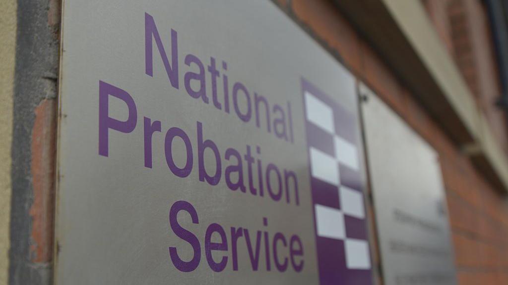 A sign with the logo of the National Probation Service written on it, attached to a red brick wall.