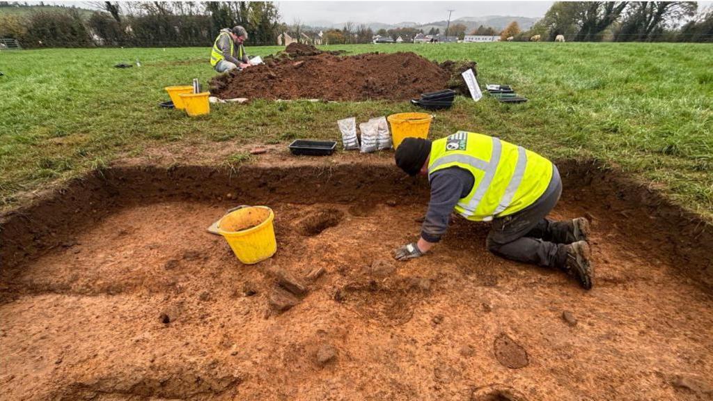 An archeological dig takes place in a field. A man in a hi-vis vest crouches in a carefully dug trench. he's wearing a hat and gloves and has a yellow bucket in front of him. in the background a man wearing similar clothing kneels in front of a pile of soil. He holds a clipboard and is surrounded by buckets.