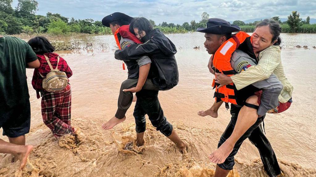 Two male police officers carry women on their backs through muddy flood waters