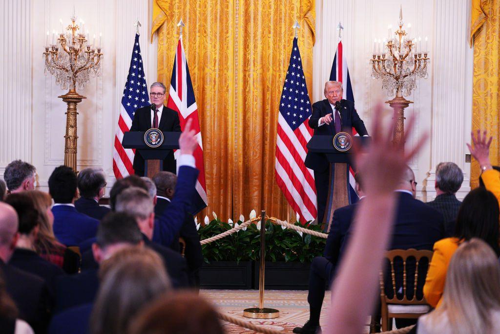 PM Starmer and President Trump stand on stage in front of flags at a press conference. Reporters are seen with hands in the air waiting to ask a question.