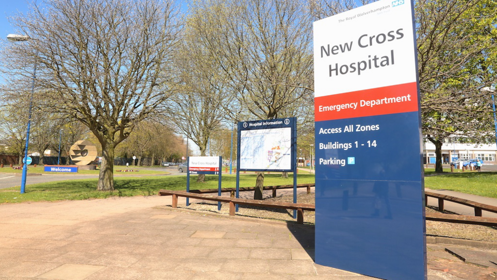 A sign that says New Cross Hospital stands on concrete with buildings behind in the distance.
