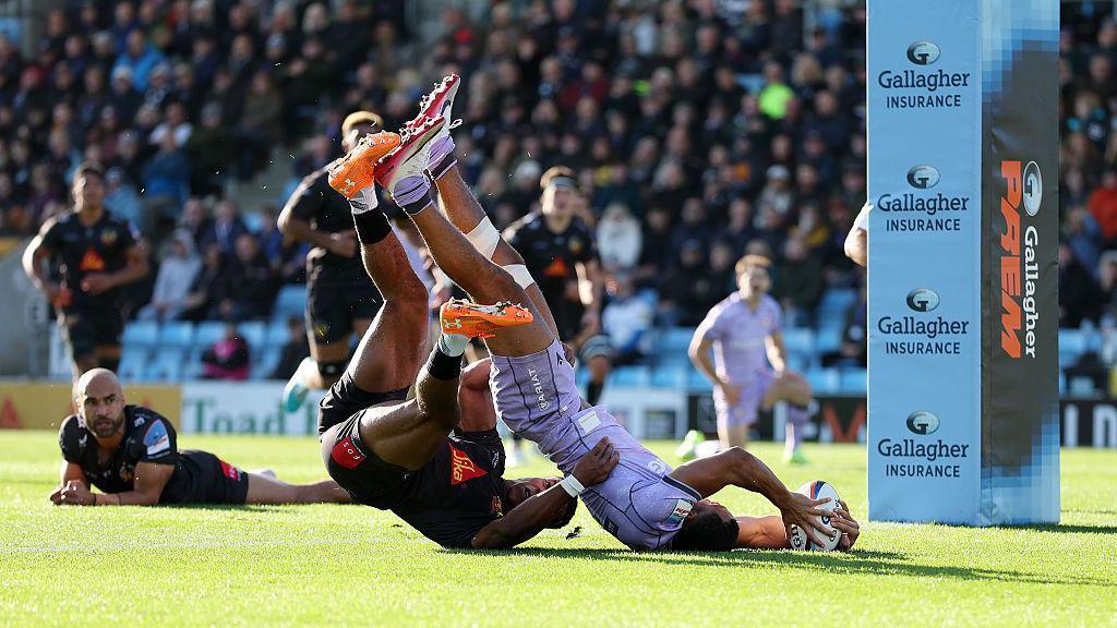 Will Joseph of Gloucester scores his side's first try against Exeter Chiefs while upside down. Exeter Chiefs' Immanuel Feyi-Waboso attempts to tackle him as other Exeter and Gloucester players watch on in the background.