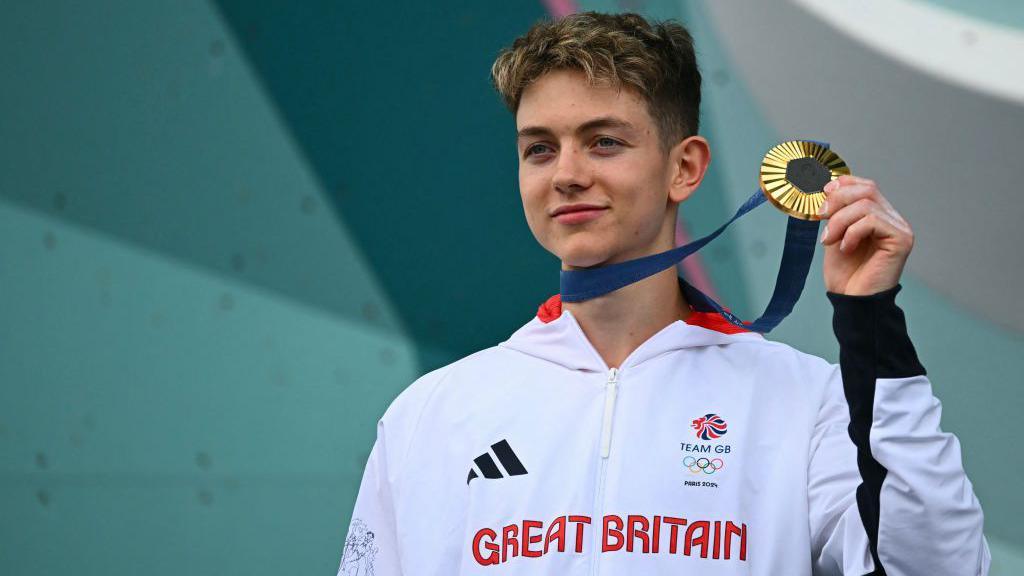 Olympic climber Toby Roberts holds up his gold medal. He's wearing an white Team GB tracksuit that reads Great Britain.
