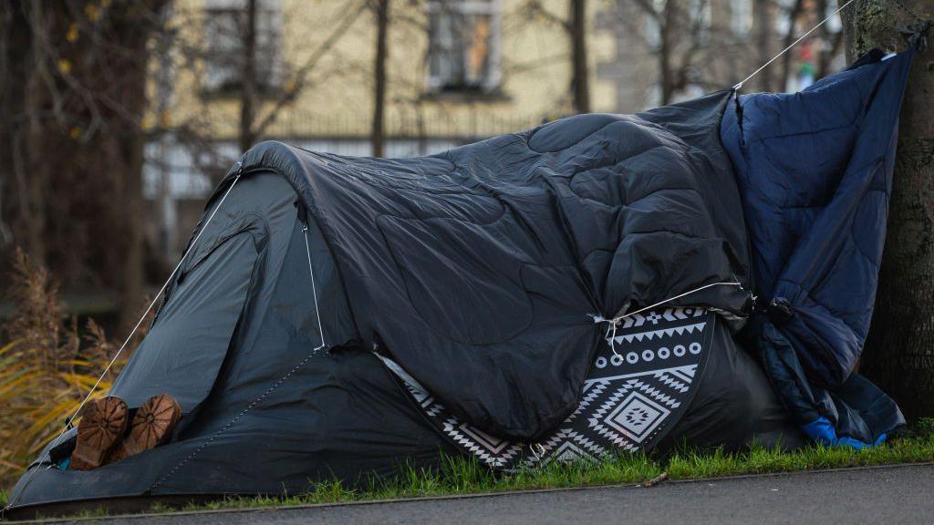 Dark blue tent on grass with a a pair of shoes sticking out of the entrance with only the soles showing. There are trees and buildings in the background.