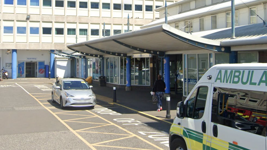 The outside of North Tees Hospital An ambulance is waiting in the yellow hatched drop off zone and a silver car is also parked. Parts of larger hospital building can be seen in the background.