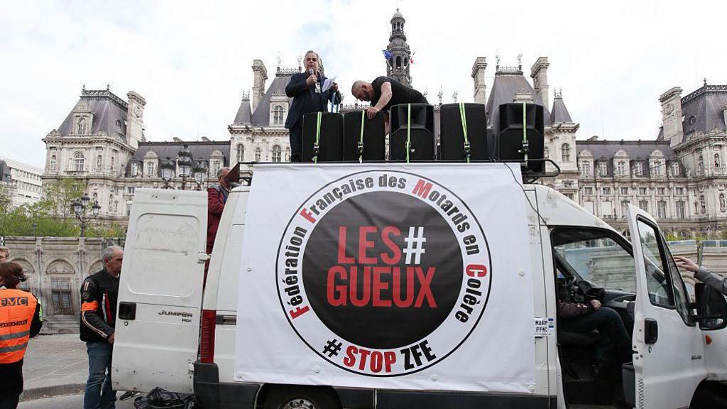 A man stands on top of a white van bearing the slogan Les #Gueux - the beggars