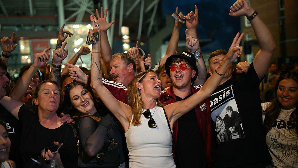 Grŵp o bobl yn sefyll yn agos at ei gilydd mewn ar ol cyngerdd Oasis yng Nghaerdydd, gyda goleuadau nos yn y cefndir. Mae sawl person yn codi eu dwylo yn yr awyr, rhai yn gwneud arwyddion heddwch ac yn dal ffonau symudol. Dillad fel crysau-t gyda logo Oasis. Yn y cefndir, gellir gweld strwythur stadiwm y Principality a rhai adeiladau brics.