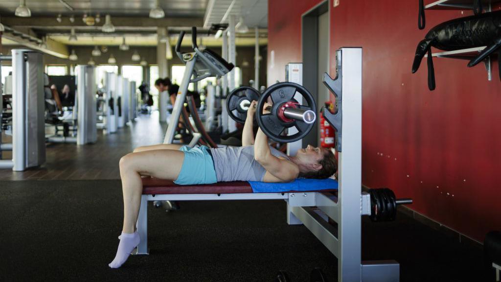 A woman lifting weights in the gym. She is lying on her back on a bench and lifting up a bar with weighted plates on each end.