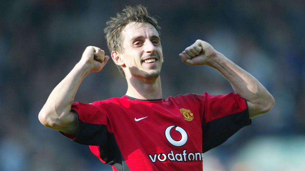 Gary Neville wearing a red Manchester United shirt holds his arms up