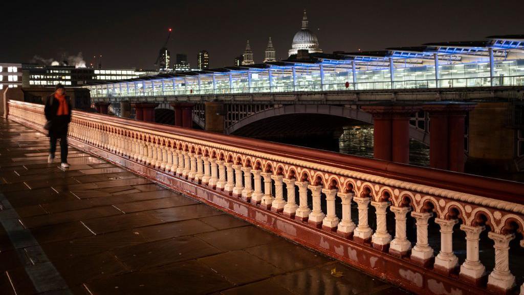 Figure walks across a wet Blackfriars Bridge, adjacent to Blackfriars Railway Bridge at night.