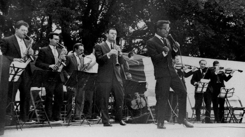 Sammy Davis Jr sing son a stage backed a by a row of men 