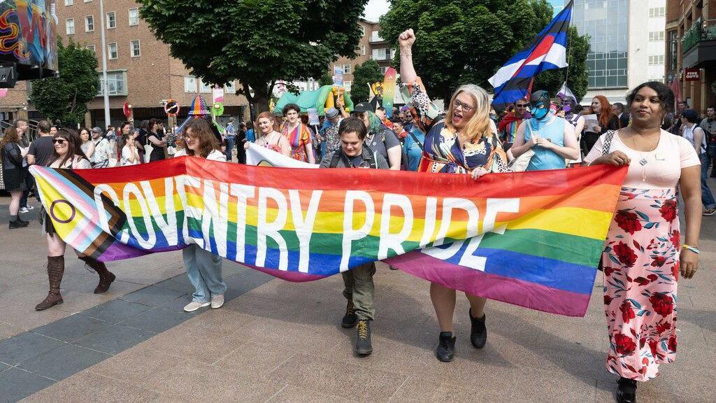 People marching in with Coventry Pride flag. One, with blonde hair, has her hand up punching the air as others hold the banner between them