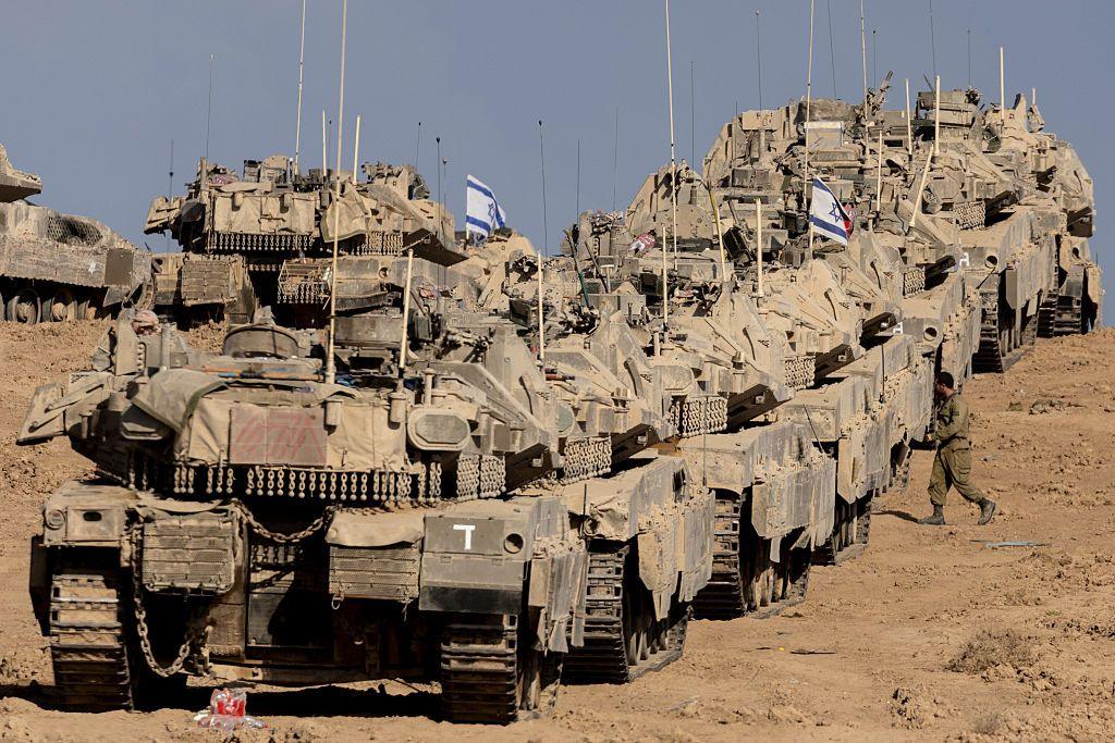 A row of at least five tanks, in a dusty barren landscape. Some have Israeli flags on them.