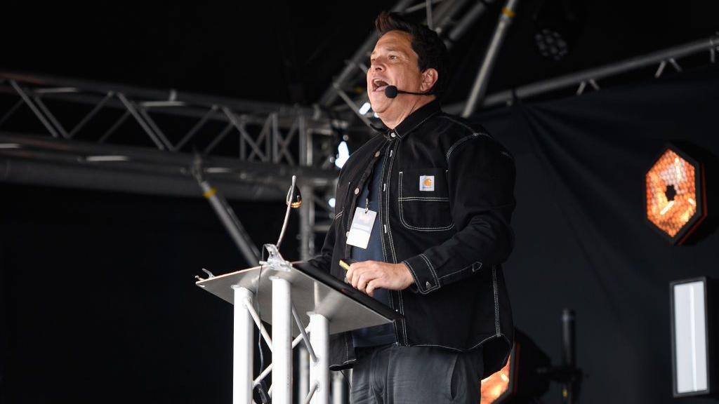 Dom Joly has dark hair and is standing at a plinth on stage. He is in a black denim jacket with dark tshirt and jeans. His mouth is open as he speaks into a microphone headset