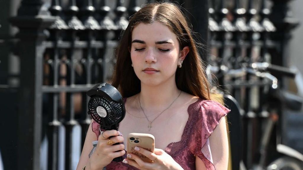 A young girl in a pink top with brown hair walks along a street carrying a portable black fan, whilst looking down at her phone in a clear case. 