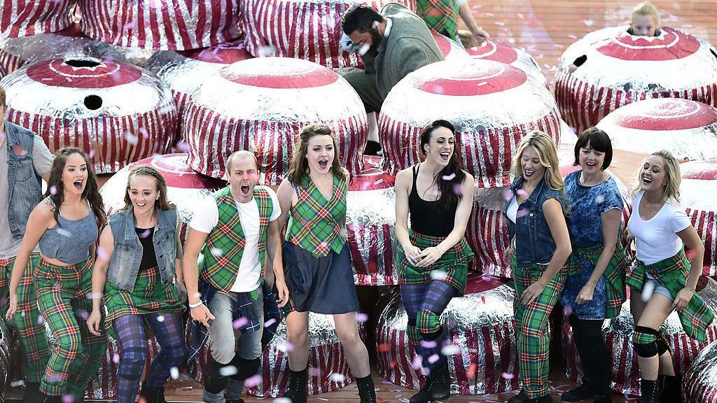 Several performers wearing tartan perform in some giant tunnocks tea cakes, wrapped in silver and red packaging