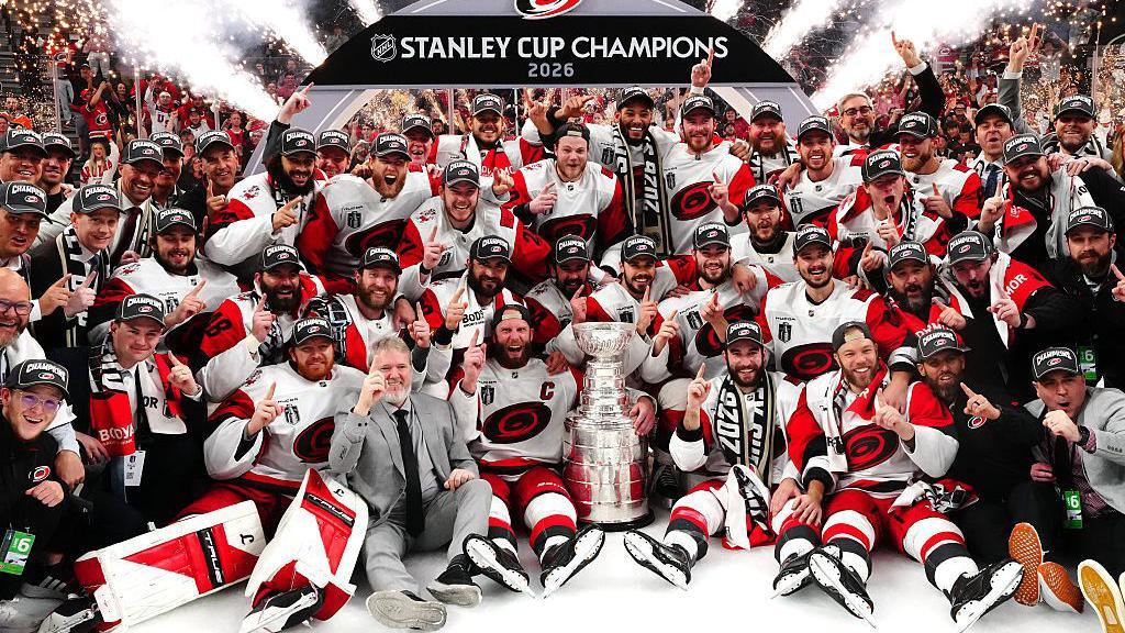The Carolina Hurricanes players, wearing white and red kits, gathered around the silver Stanley Cup trophy