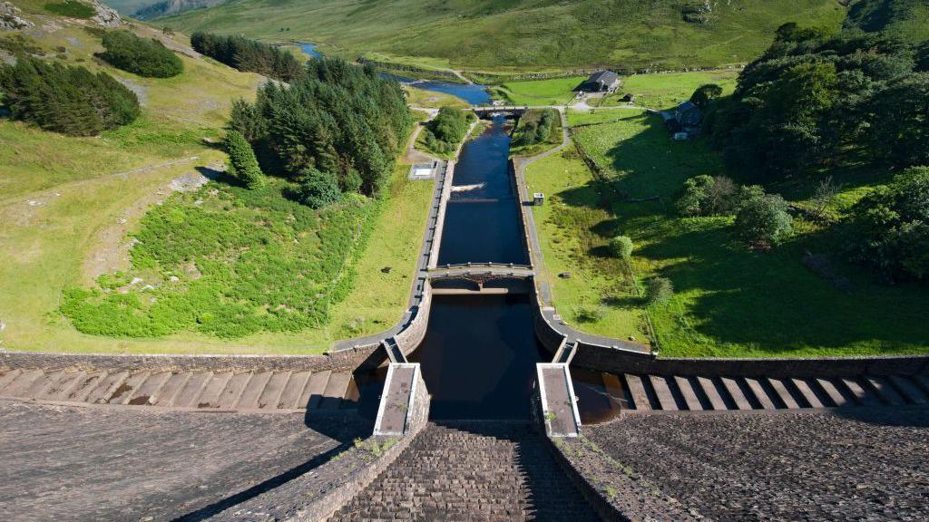 water overflow from the dam of the Claerwen Reservoir with green hills and trees to the sides