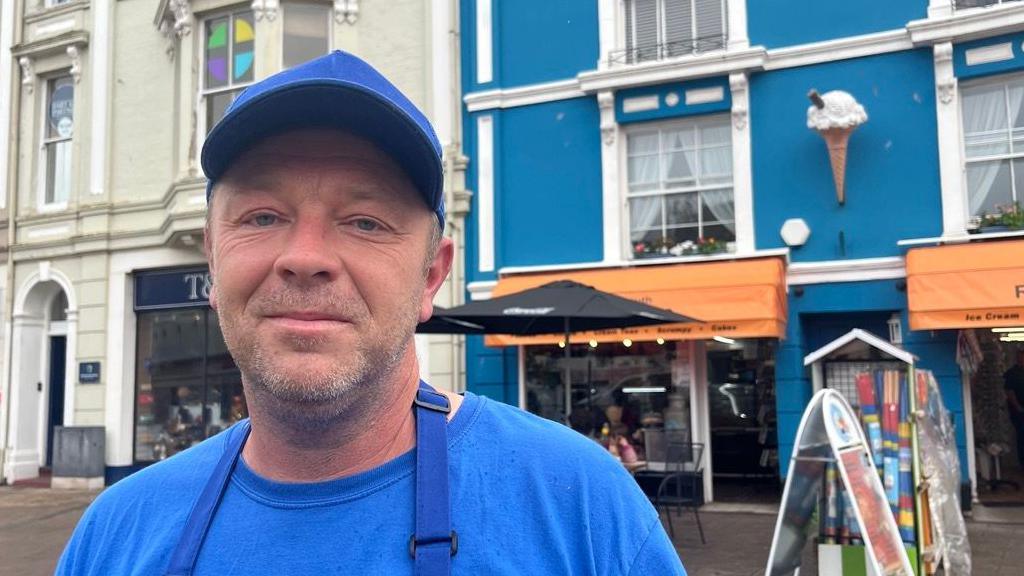 Clive Wetton wearing a blue cap, a blue T-shirt and a blue apron. He is standing in front of the Teignmouth Bake House with its orange awning and a black parasol at the front of the cafe.