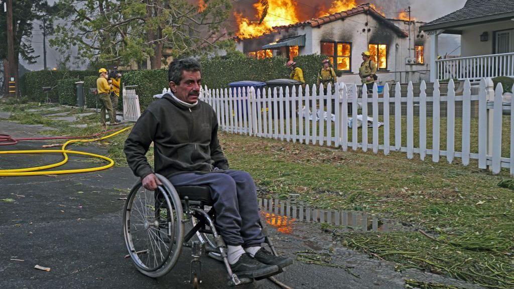 A man in a wheelchair goes past a house on fire from the Eaton Fire in the Altadena neighborhood on January 08, 2025 in PASADENA, CALIFORNIA. 