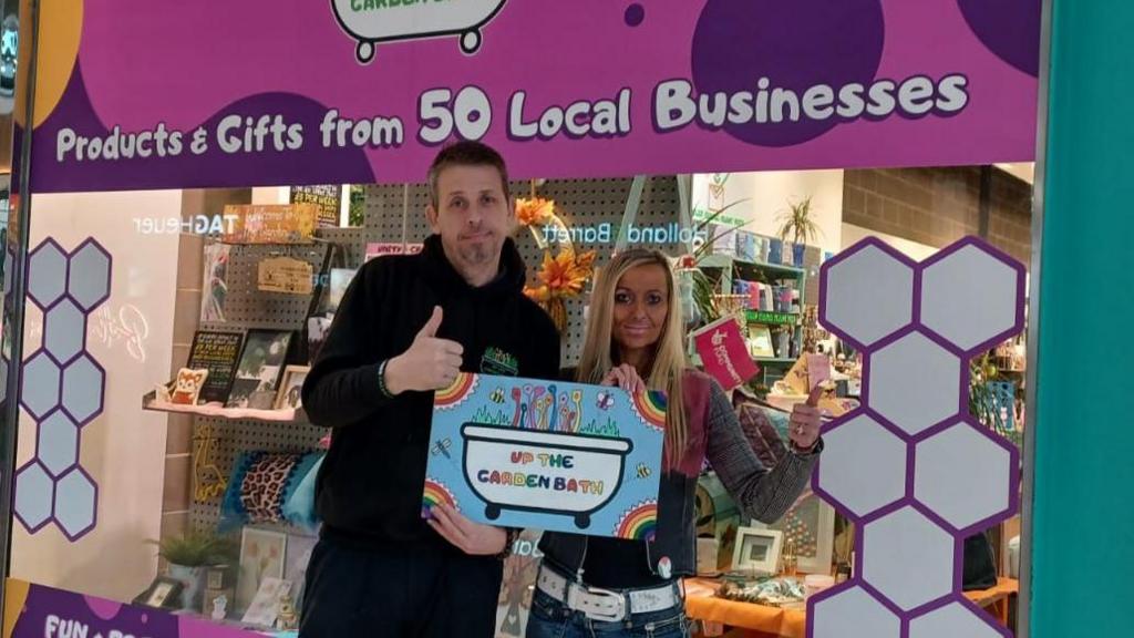 Dave and Kez are stood in front of the Unity shop window holding a "Created by Up the Garden Bath" sign in their hands. Dave is wearing a black hoodie with the Garden Bath logo on the front right and is giving a thumbs up to the camera. Kez, who has long blonde hair,  is doing the same thumbs up gesture. Behind them in the shop window a sign reads "Products and gifts from 50 local businesses". 