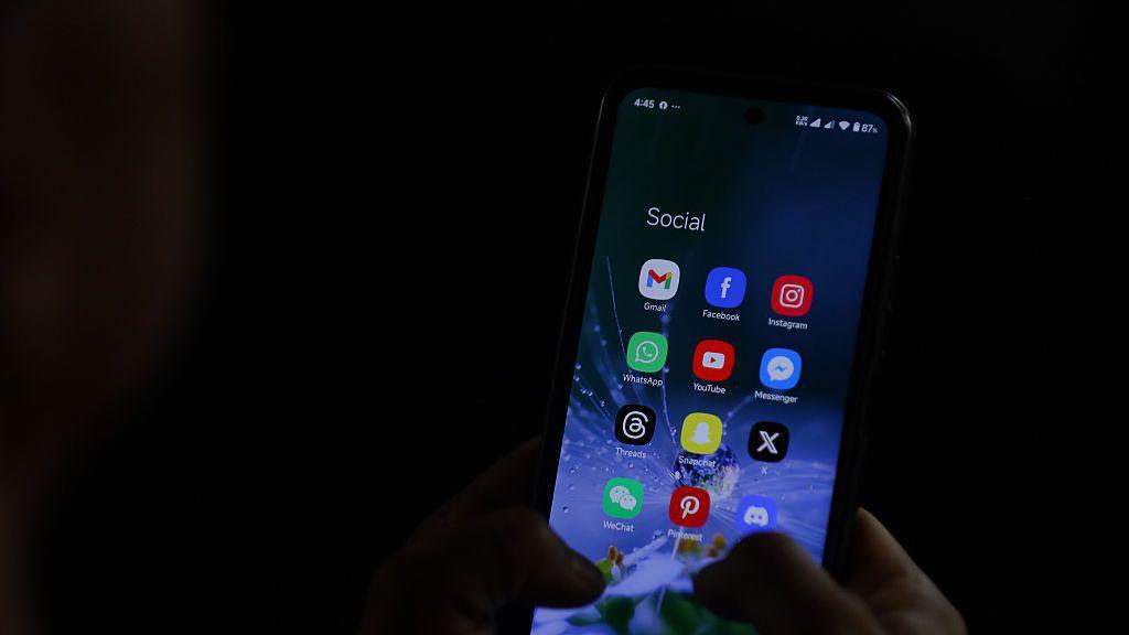 A smartphone being used in a dark room. A person is holding it in their hand. The screen has been opened to a page that says "Social" and contains social media apps including X, Snapchat, Instagram, Facebook and Threads.