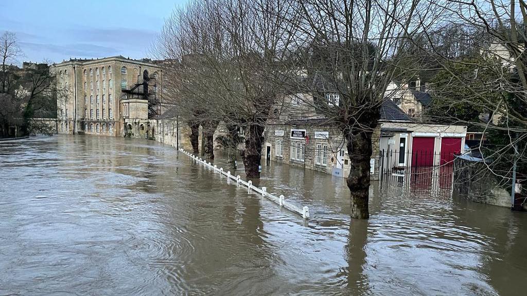 A shot of Bradford-on-Avon during flooding - bottoms of doors and buldings submerged, water almost up to the top of a fence, trees surrounded by brown rover water.