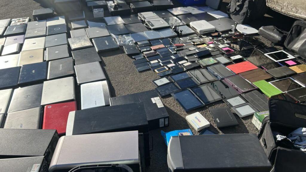 A wide array of IT devices lined up on a street next to a van.