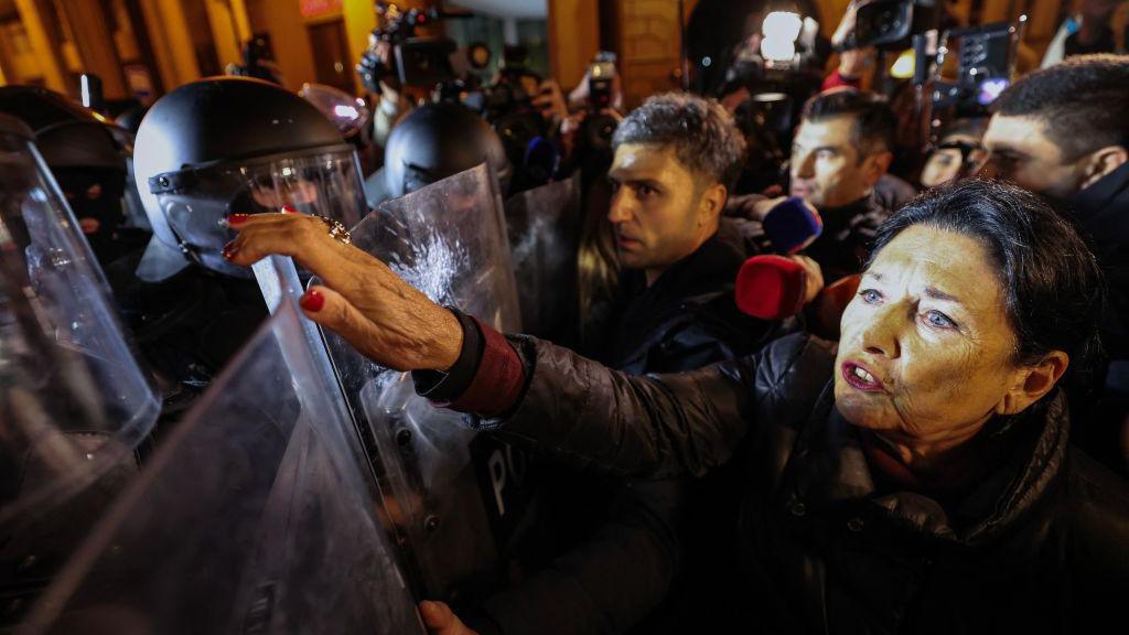 Georgia's President Salome Zourabishvili, a woman with black hair, stands directly in front of riot police, reaching up over a shield towards a masked officer.