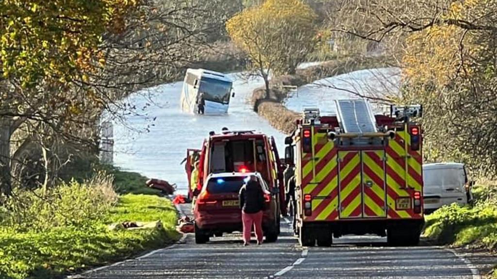 A bus tilting in flood water in the distance, with fire and rescue vehicles closer at the edge of the water