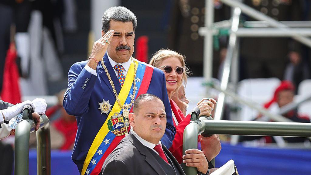 President of Venezuela Nicolás Maduro, wearing a suit and the presidential sash, salutes as he arrives for a military parade as part of 214th anniversary of Venezuela's independence celebrations on July 5, 2025 in Caracas, Venezuela. A man stands in front of him and Maduro's wife, Cilia Flores, smiles behind him.