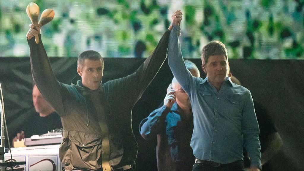 Two men raise each others hands. The man on the left is wearing a dark green raincoat and holds two shakers in his right hand. The man on the right wears a blue denim shirt, top button un-done, They both have short brown hair. Behind them are several men who stand in front of a screen displaying green patterns. 