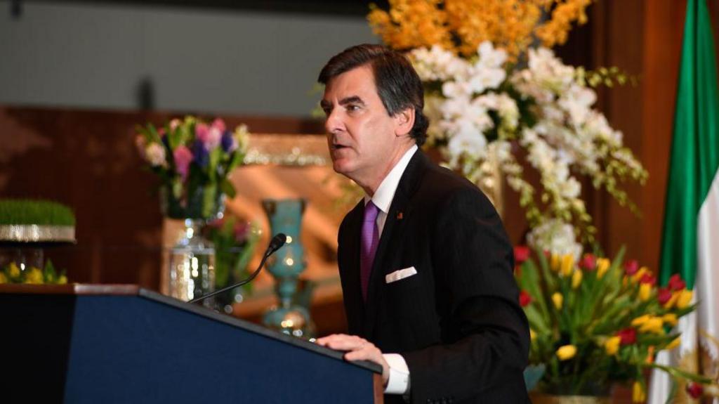 Man wearing a suit and tie standing at a podium with his hand on the podium speaking into a microphone. In the background several bunches of flowers.